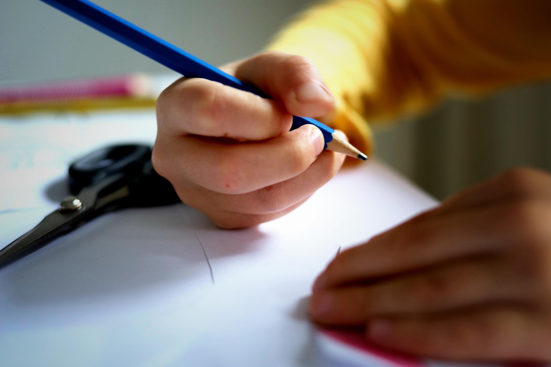 Nahaufnahme einer Kinderhand, die einen blauen Bleistift hält und auf weißem Papier zeichnet oder schreibt. Auf dem Tisch ist eine Schere zu sehen, und das Kind trägt ein gelbes langärmeliges Hemd.