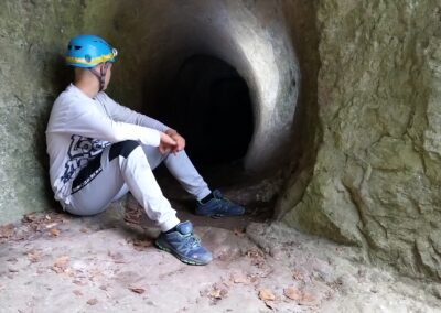 Eine Person in sportlicher Kleidung und mit blauem Helm sitzt am Eingang einer dunklen Höhle und schaut hinein. Die Wände der Höhle sind rau und felsig, und trockenes Laub liegt auf dem Boden verstreut.