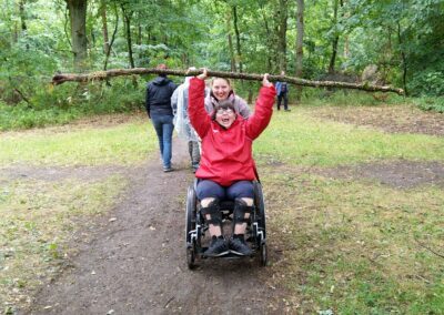 Eine lächelnde Person in einem Rollstuhl hält mit Hilfe einer stehenden Frau hinter ihr in einem grünen, bewaldeten Park einen großen Ast über ihren Kopf. Zwei weitere Personen gehen im Hintergrund.