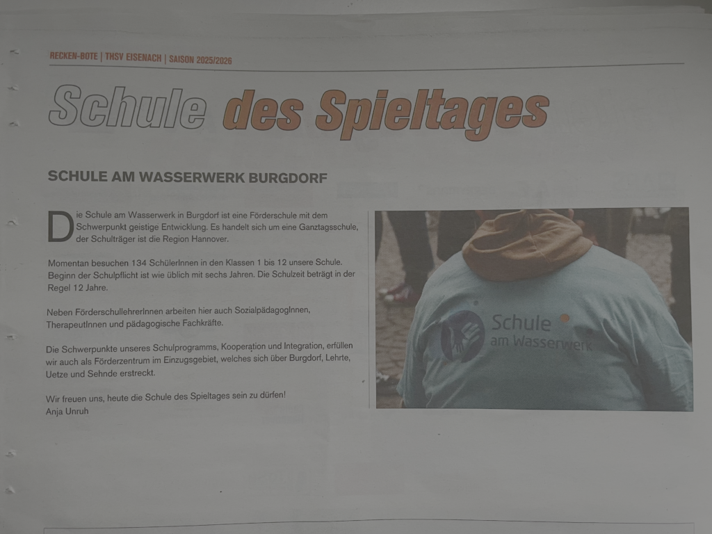 Auf einer Zeitschriftenseite ist ein Artikel über die "Schule des Spieltages" mit einem Foto einer Person in einer hellblauen Jacke abgebildet. Die Jacke ist auf der Rückseite mit dem Logo und dem Text "Schule am Wasserwerk" bedruckt.