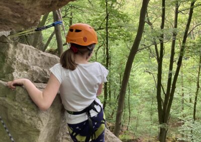 Eine Person, die einen Helm und einen Klettergurt trägt, klettert in einem Wald auf einen felsigen Abhang. Sie hält sich mit einer Hand am Felsen fest und blickt auf grüne Bäume, über denen eine Kletterausrüstung zu sehen ist.