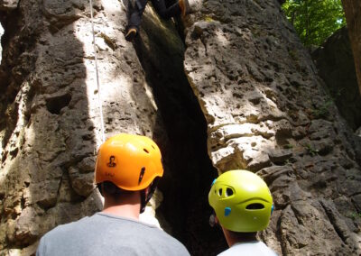Zwei Personen mit Helmen beobachten einen Kletterer, der eine felsige Klippe im Freien erklimmt. Das Sonnenlicht fällt durch die Bäume über ihnen und beleuchtet die strukturierte Felsoberfläche und den Kletterer in der Nähe des Gipfels.
