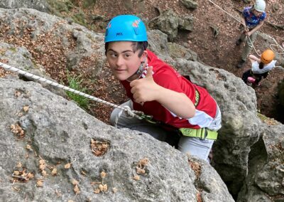 Ein Junge mit blauem Helm und rotem Hemd klettert auf einen Felsen und zeigt mit dem Daumen nach oben in die Kamera. Zwei andere Kletterer mit Helmen sind unten auf dem Boden in einem Waldgebiet zu sehen.