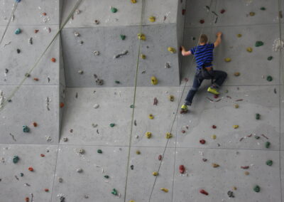 Eine Person in einem blau gestreiften Hemd und Jeans klettert an einer Indoor-Kletterwand mit bunten Griffen und Sicherungsseilen.