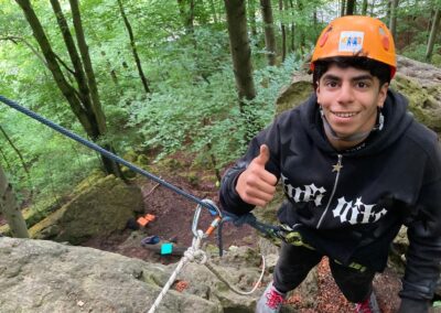 Ein Jugendlicher, der einen orangefarbenen Helm und einen schwarzen Kapuzenpulli trägt, zeigt den Daumen nach oben, während er mit Kletterseilen und Klettergurten gesichert auf Felsen in einem grünen Wald klettert.