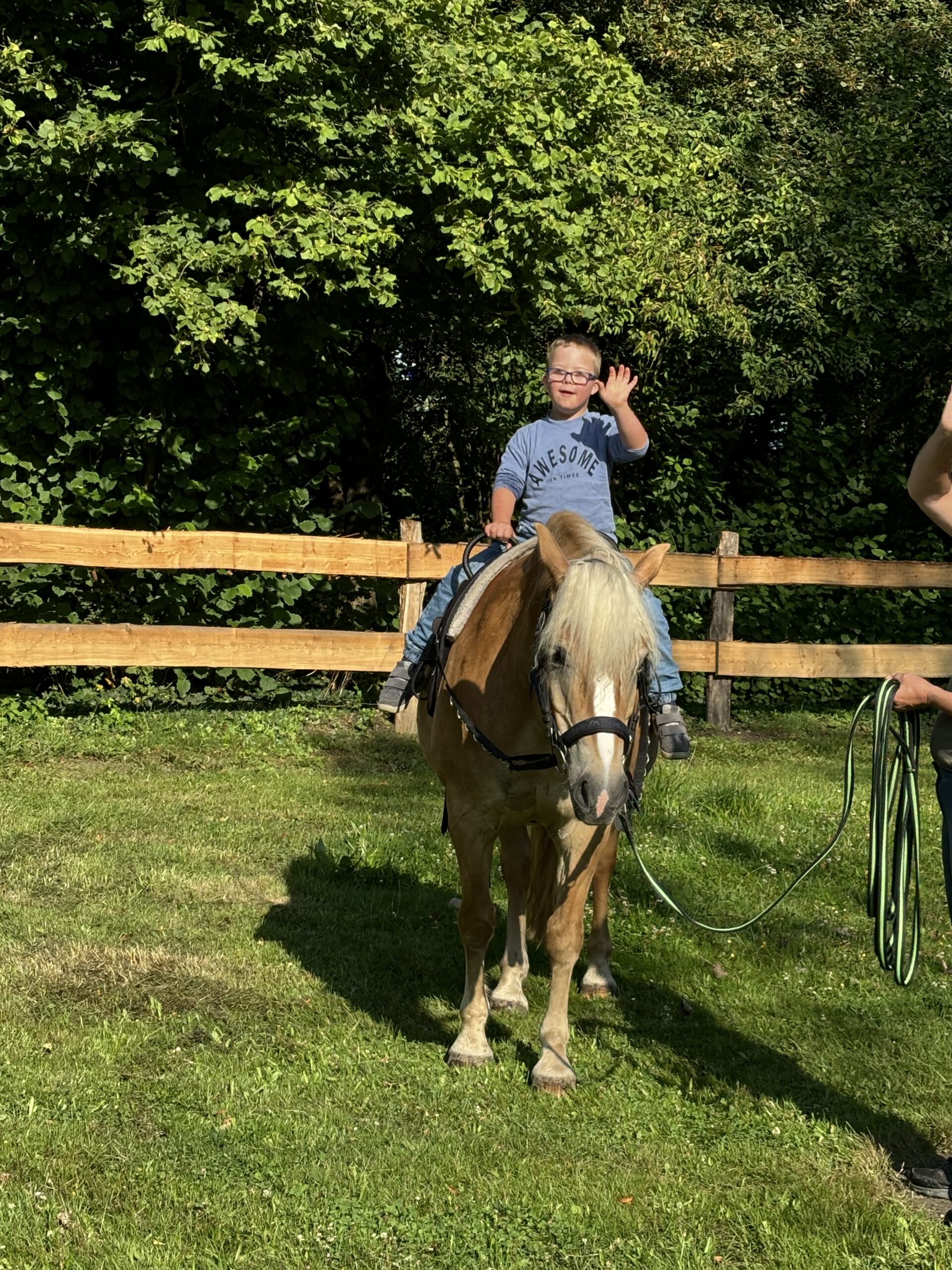 Reiten Therapie Ein kleiner Junge mit Brille und grauem Awesome-T-Shirt sitzt auf einem hellbraunen Pferd und lächelt und winkt. Das Pferd wird von einem Erwachsenen mit grünen Zügeln geführt. Sie befinden sich draußen in der Nähe eines Holzzauns und von Bäumen.