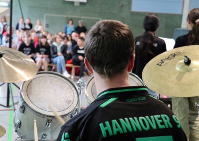 Ein Schlagzeuger im Trikot von Hannover 96 spielt vor einem Publikum in einer Sporthalle, wobei im Vordergrund ein Schlagzeug und Becken zu sehen sind und im Hintergrund eine Menschenmenge sitzt.