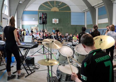 Eine Band tritt auf der Bühne einer Sporthalle auf, mit einem Schlagzeuger im Vordergrund und einem Keyboarder auf der linken Seite. Das Publikum sitzt auf Bänken und beobachtet den Auftritt. Die Szene befindet sich in einem Innenraum mit hohen, gewölbten Decken.