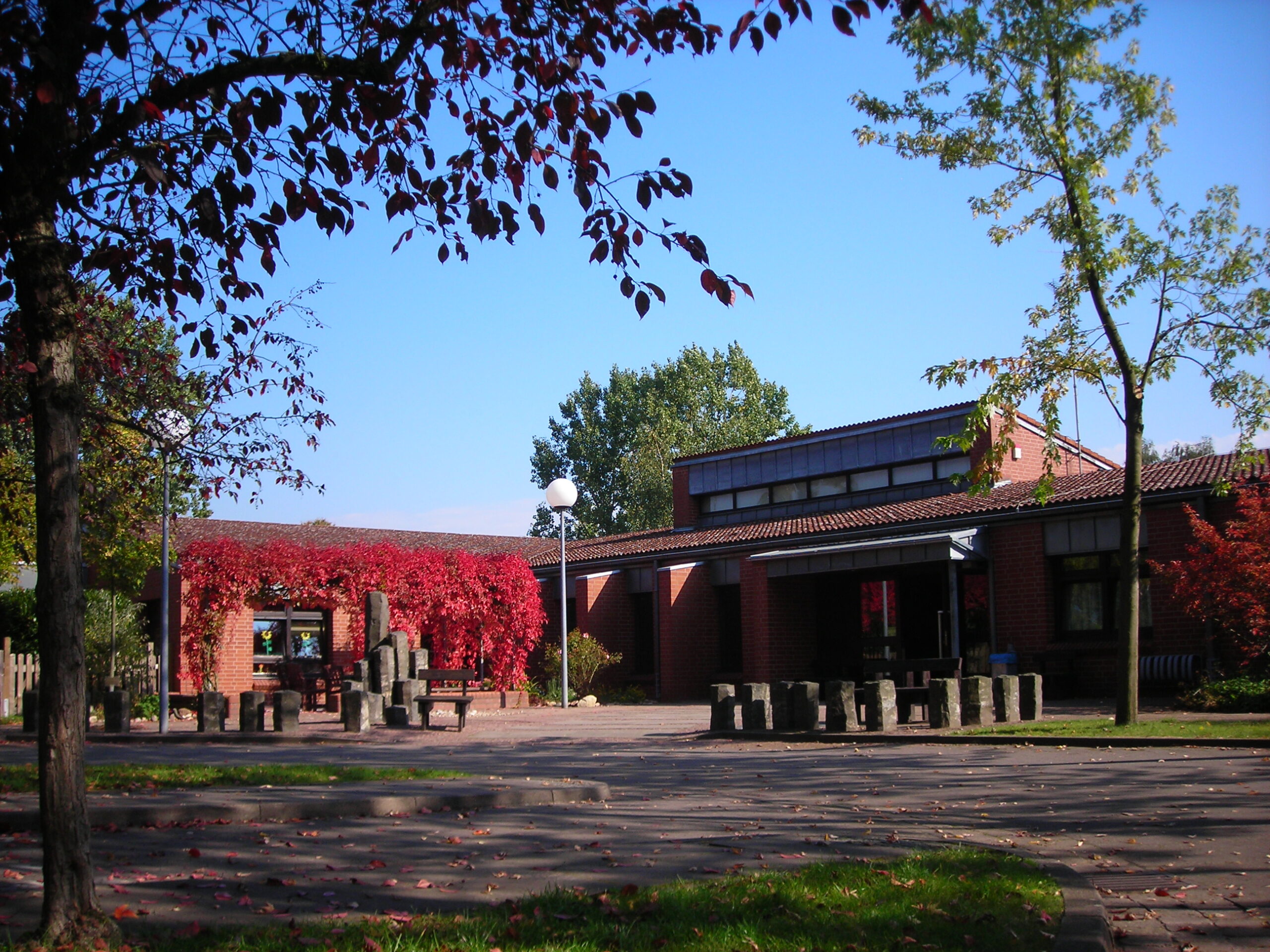 Ein Backsteingebäude mit großen Fenstern und einem überdachten Gang steht hinter einem von Bäumen beschatteten Innenhof. Rotes Herbstlaub bedeckt einen Teil des Weges, und in dem offenen, sonnenbeschienenen Bereich sind runde Steinsitze aufgestellt.
