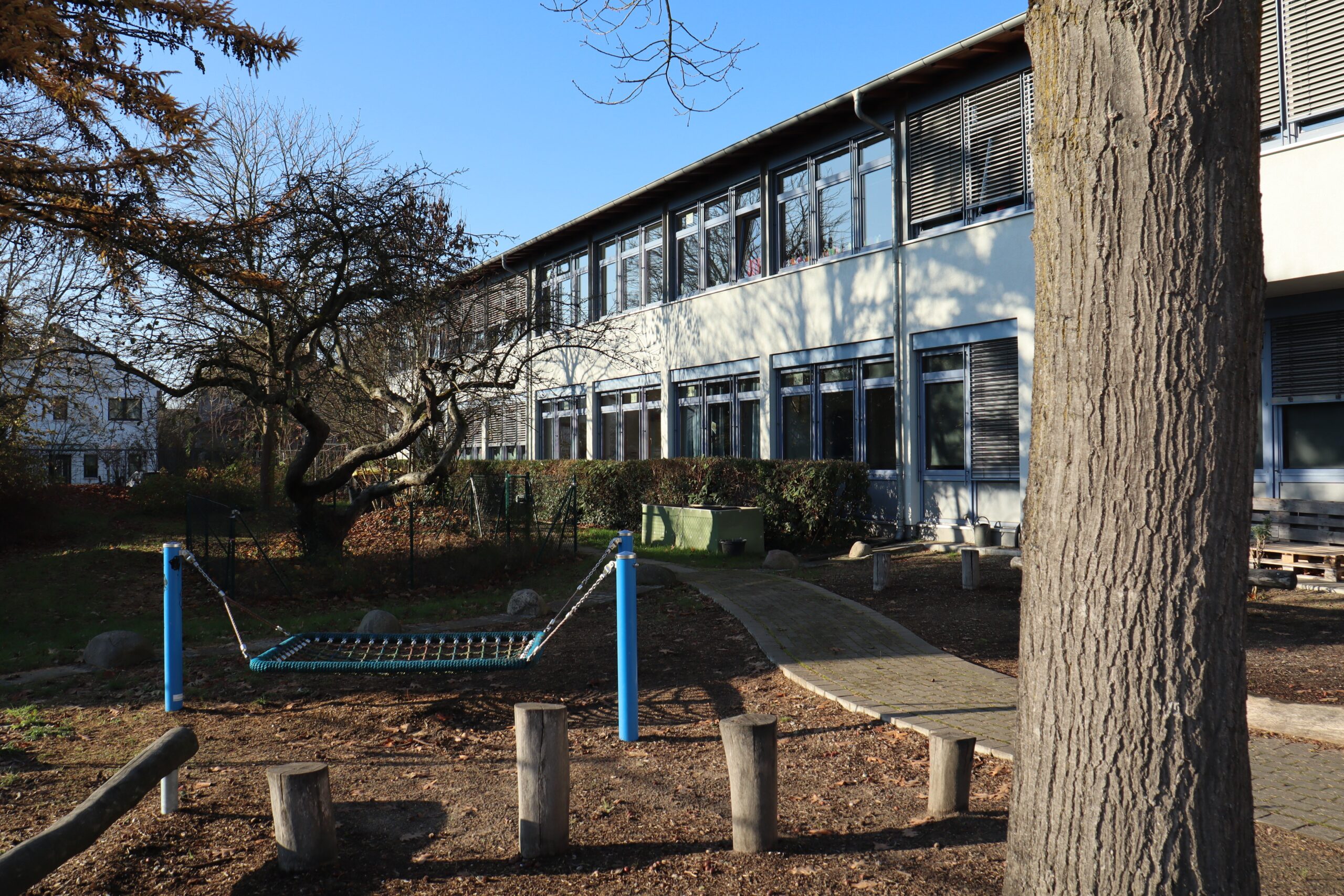 Ein Schulgebäude mit großen Fenstern ist unter einem klaren blauen Himmel zu sehen. Im Vordergrund befinden sich eine Hängemattenschaukel, Baumstämme und ein gepflasterter Weg, der sich durch den Außenbereich schlängelt.