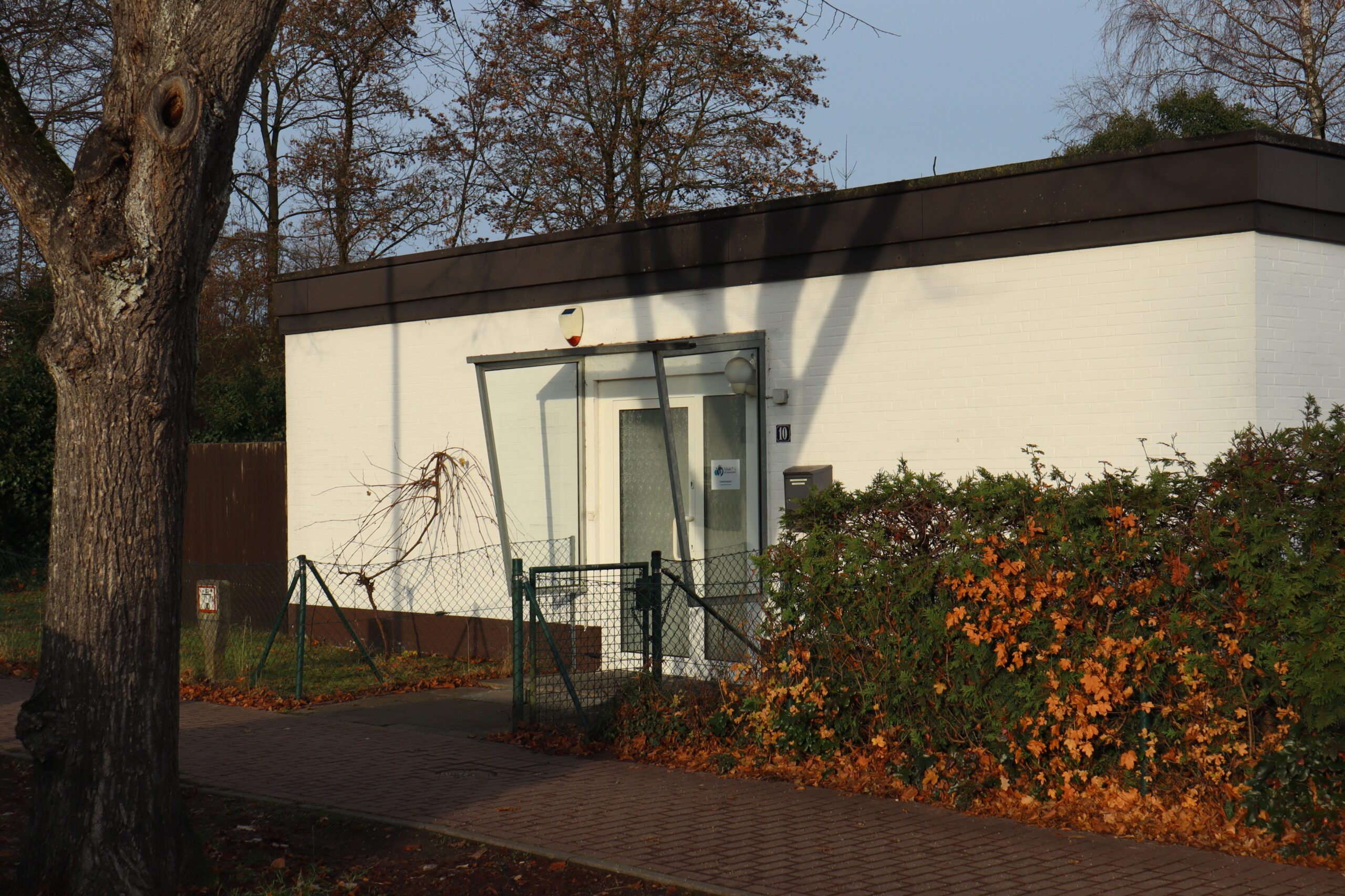 Ein kleines weißes Gebäude mit Flachdach und gläserner Eingangstür steht hinter einem niedrigen Maschendrahtzaun. Herbstlaub bedeckt den Boden und die Sträucher, und kahle Bäume werfen Schatten auf die Mauer.