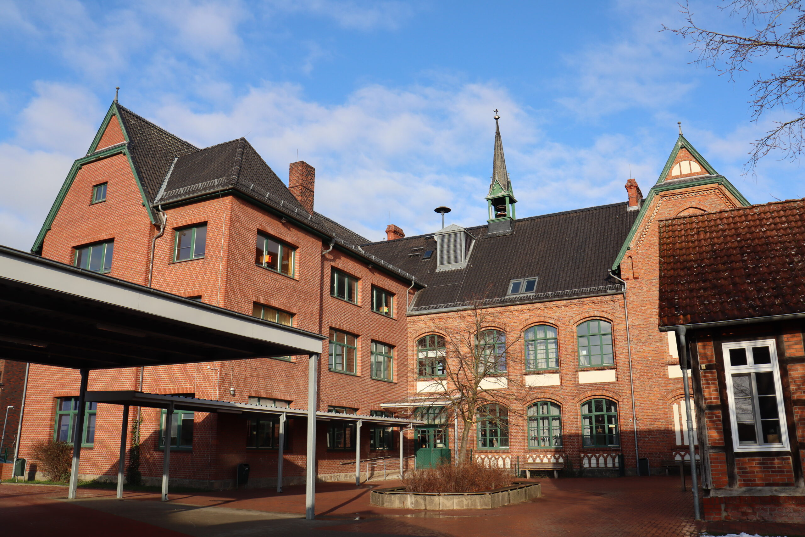 Ein Schulgebäude aus rotem Backstein mit Satteldächern, Dachgauben und einem kleinen Kirchturm, dargestellt unter einem teilweise bewölkten blauen Himmel. Der Innenhof hat gepflasterte Gehwege und einen überdachten Eingangsbereich.