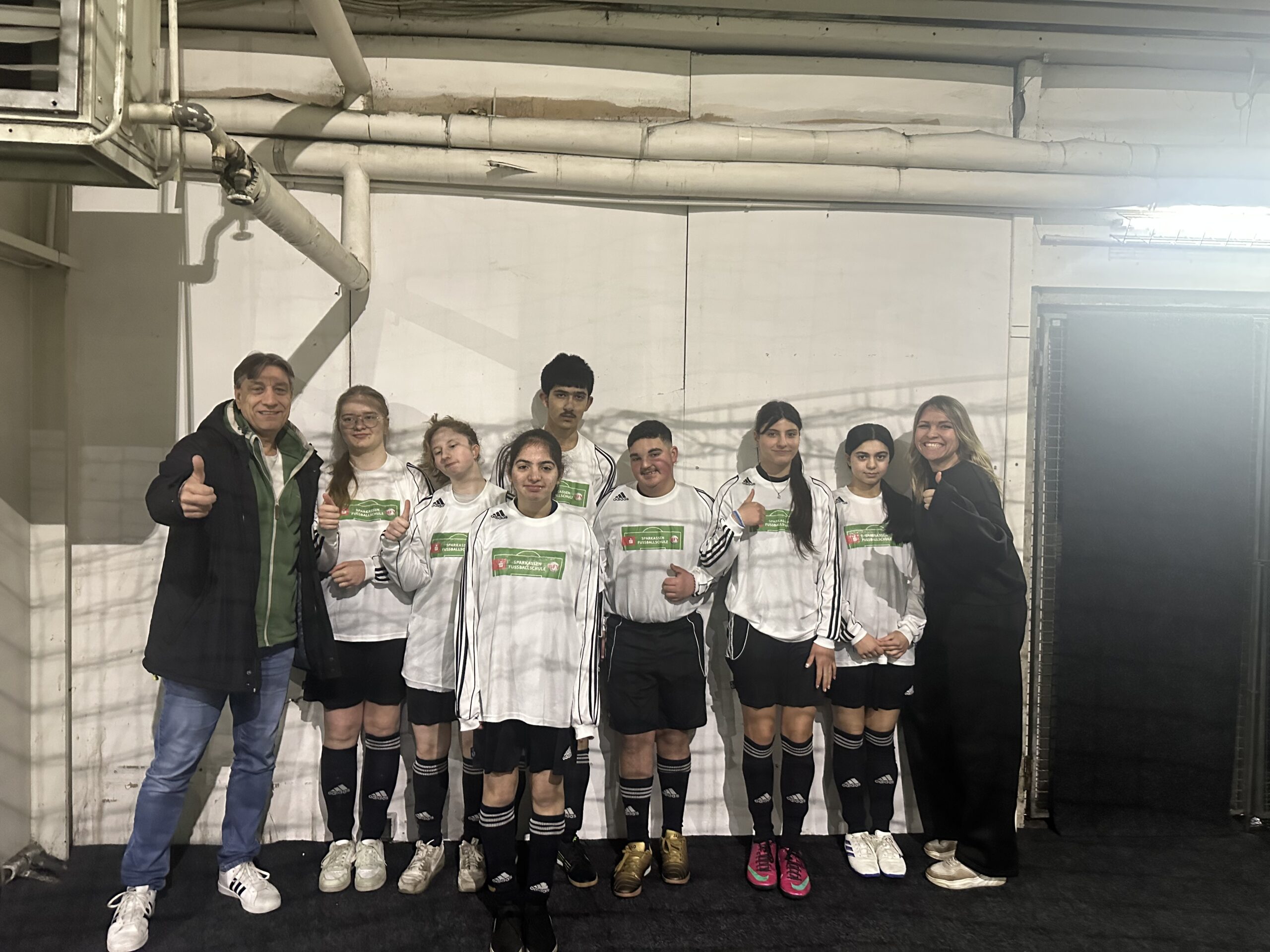 Eine Gruppe von sieben jungen Fußballspielern in weißen Trikots und zwei Erwachsene stehen in einer Halle, lächeln und zeigen die Daumen nach oben und posieren für ein Mannschaftsfoto vor einer weißen Wand.