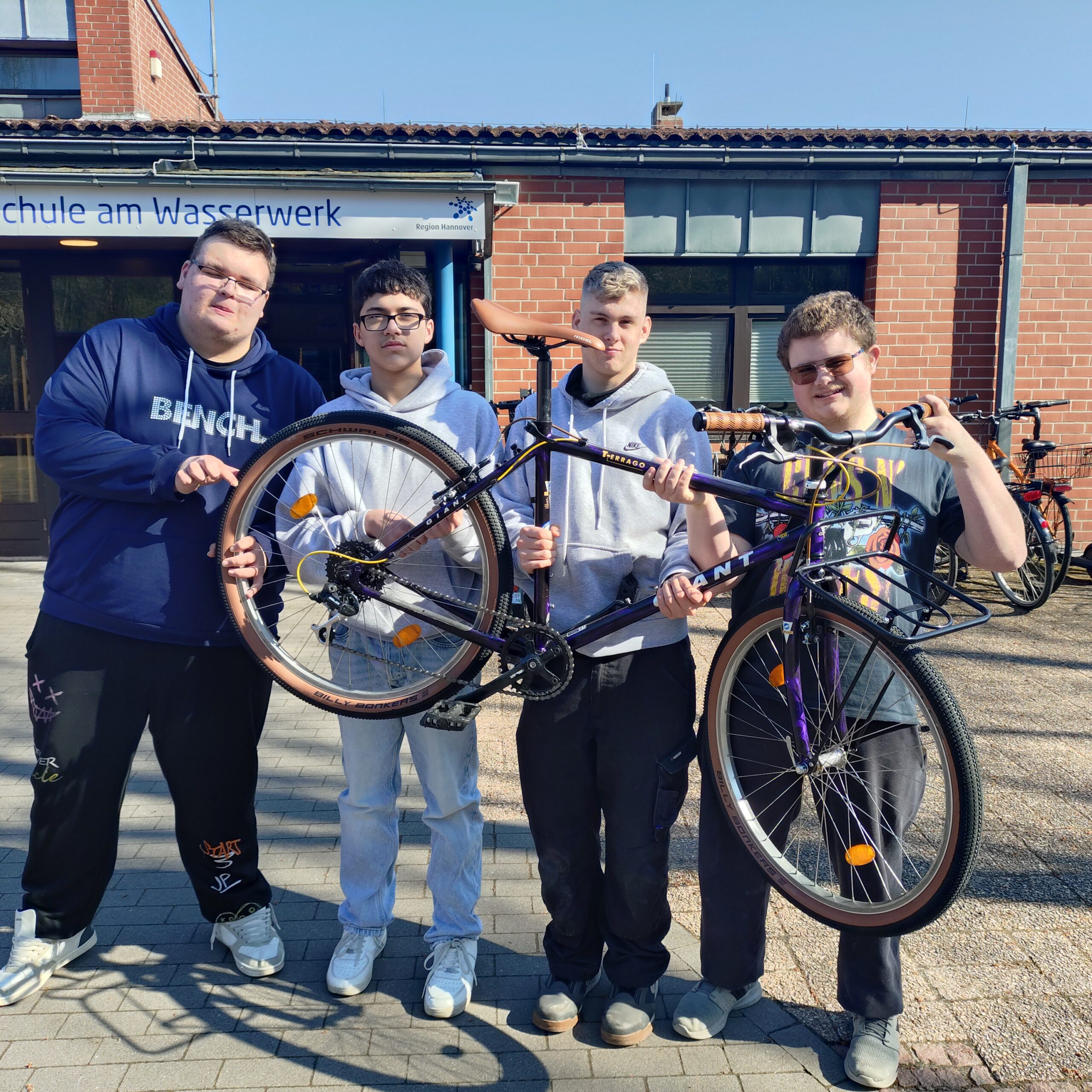 Fahrrad-AG1 Vier junge Männer stehen vor einem Backsteingebäude und lächeln in die Kamera, während sie gemeinsam ein Fahrrad hochhalten. An dem Gebäude hinter ihnen ist ein Schild in deutscher Sprache zu sehen. Es ist ein sonniger Tag.