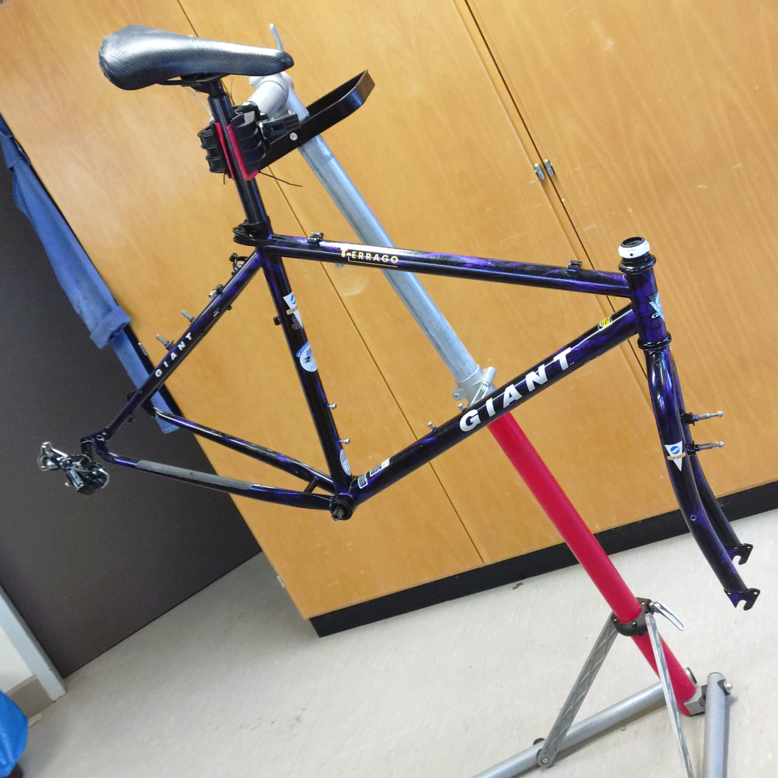 Fahrrad-AG2 Ein lilafarbener und blauer Giant-Rennradrahmen ist auf einem Reparaturständer in einem Raum mit hellem Bodenbelag und Holzschränken im Hintergrund montiert. Dem Rahmen fehlen Räder, Lenker und eine Kette.