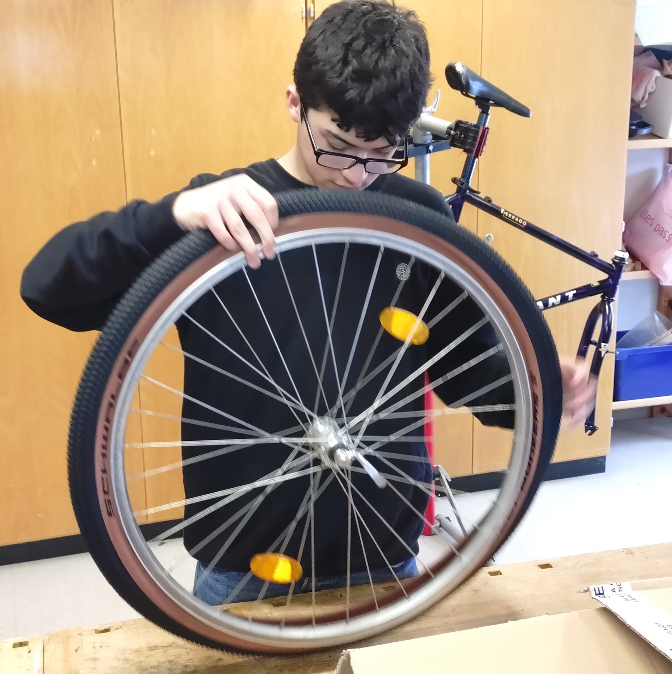 Fahrrad-AG3 Eine Person mit Brille und schwarzem Sweatshirt hält ein Fahrradrad und scheint es zu reparieren oder zu überprüfen. Im Hintergrund ist ein teilweise montierter Fahrradrahmen auf einem Holztisch zu sehen.