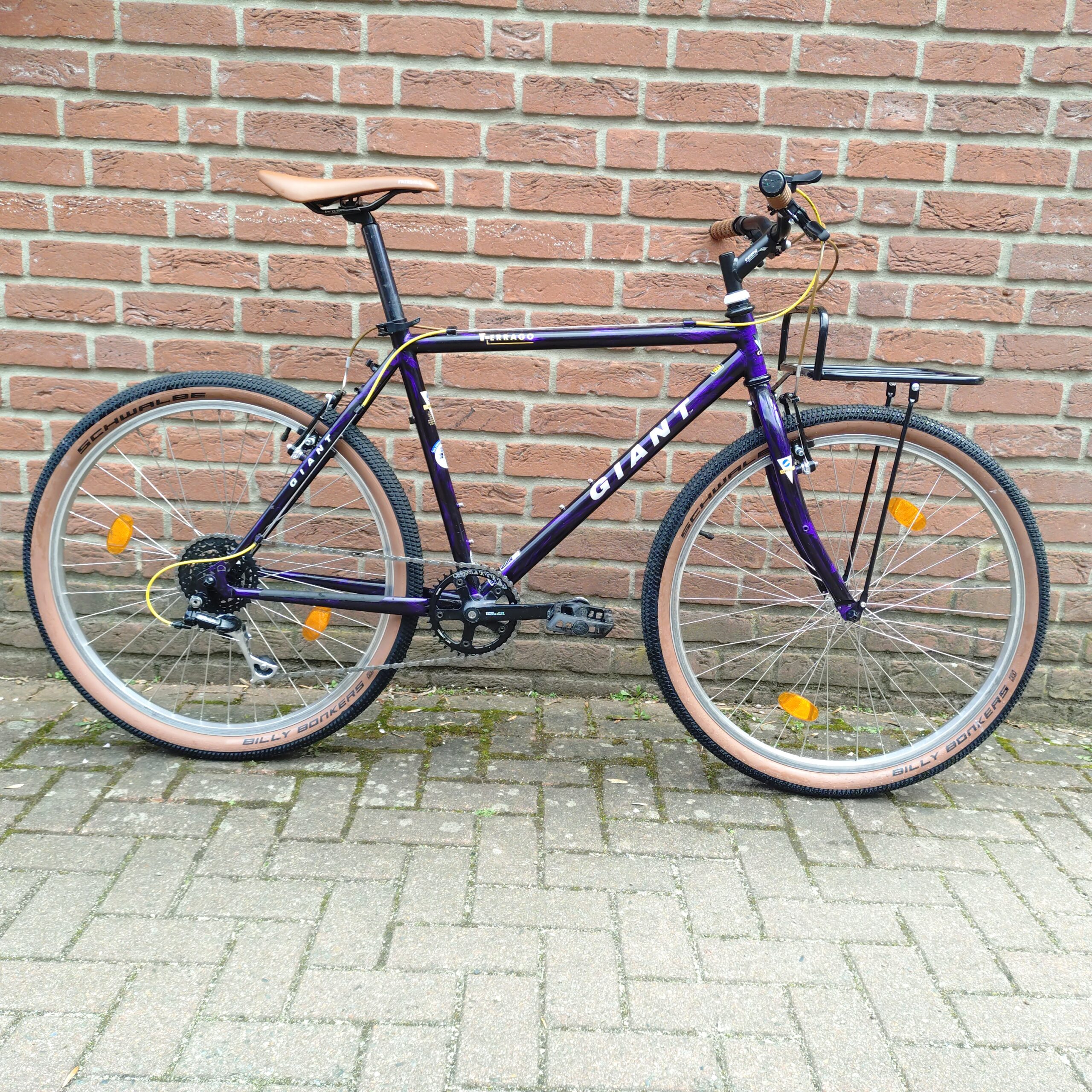 Ein lilafarbenes Giant-Mountainbike mit hellbraunen Reifen ist auf einem gepflasterten Gehweg vor einer roten Backsteinmauer geparkt. Es hat einen braunen Sitz und braune Lenkergriffe sowie orangefarbene Reflektoren an den Rädern.