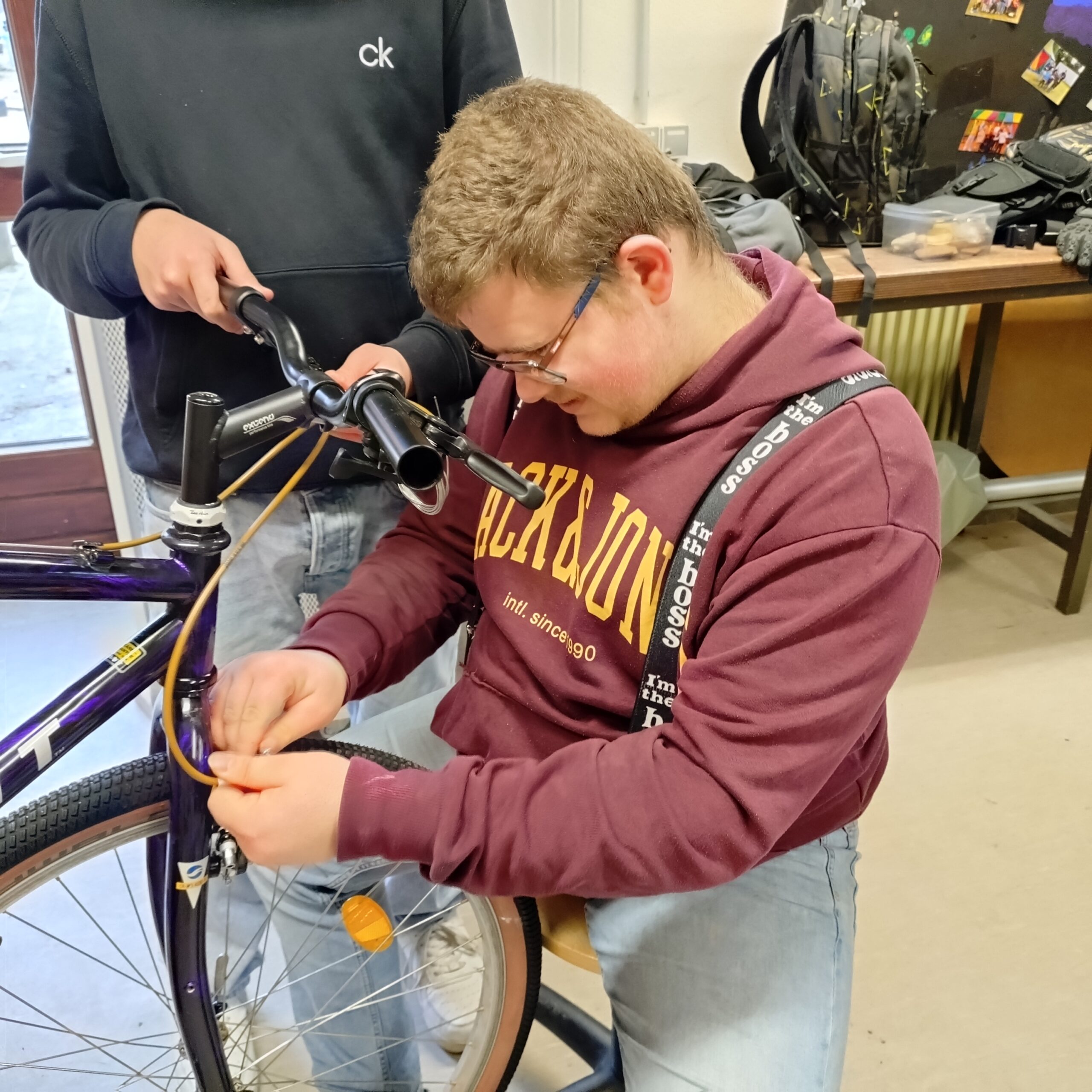 Fahrrad-AG86 Zwei Personen arbeiten zusammen, um ein Fahrrad zu reparieren. Eine Person in einem kastanienbraunen Kapuzenpullover und blauen Jeans konzentriert sich darauf, das Vorderrad des Fahrrads einzustellen, während eine andere daneben steht und den Lenker hält.