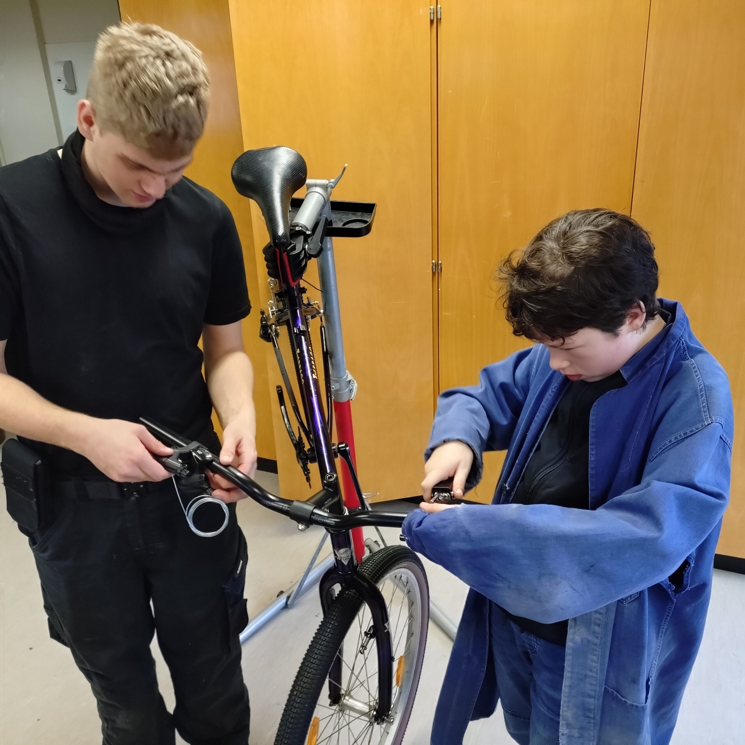 Fahrradd-AG5 Zwei Personen reparieren in einem Haus ein Fahrrad. Eine Person hält den Lenker fest, während die andere eine Schraube in der Nähe des Rades anzieht. Beide sind auf ihre Arbeit konzentriert, und im Hintergrund sind Holzschränke zu sehen.