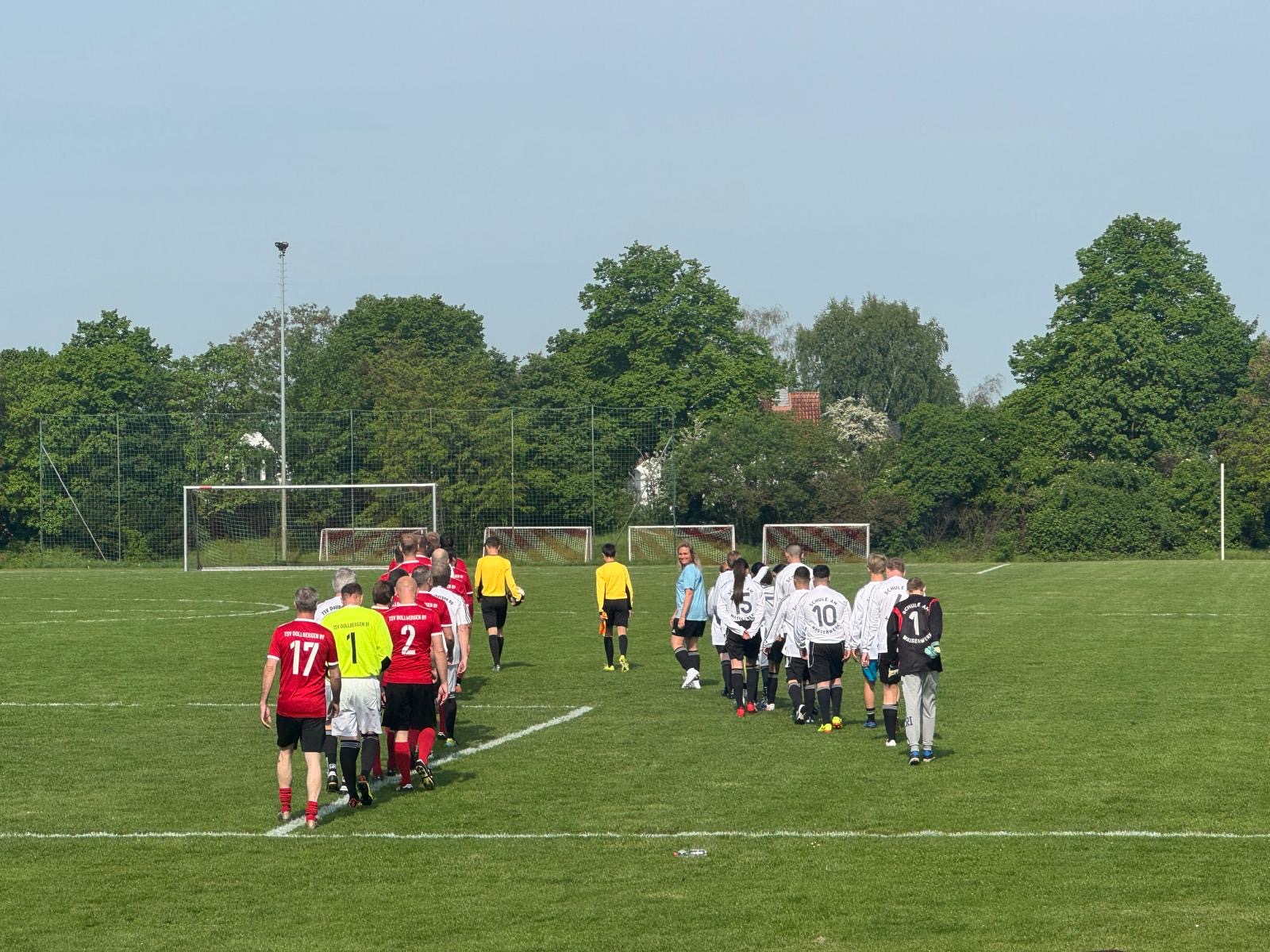 Zwei Fußballmannschaften, eine in Rot und eine in Weiß, gehen vor einem Spiel auf ein grünes Feld zu den Schiedsrichtern. Im Hintergrund sind Bäume und Torpfosten zu sehen, und der Himmel ist klar.