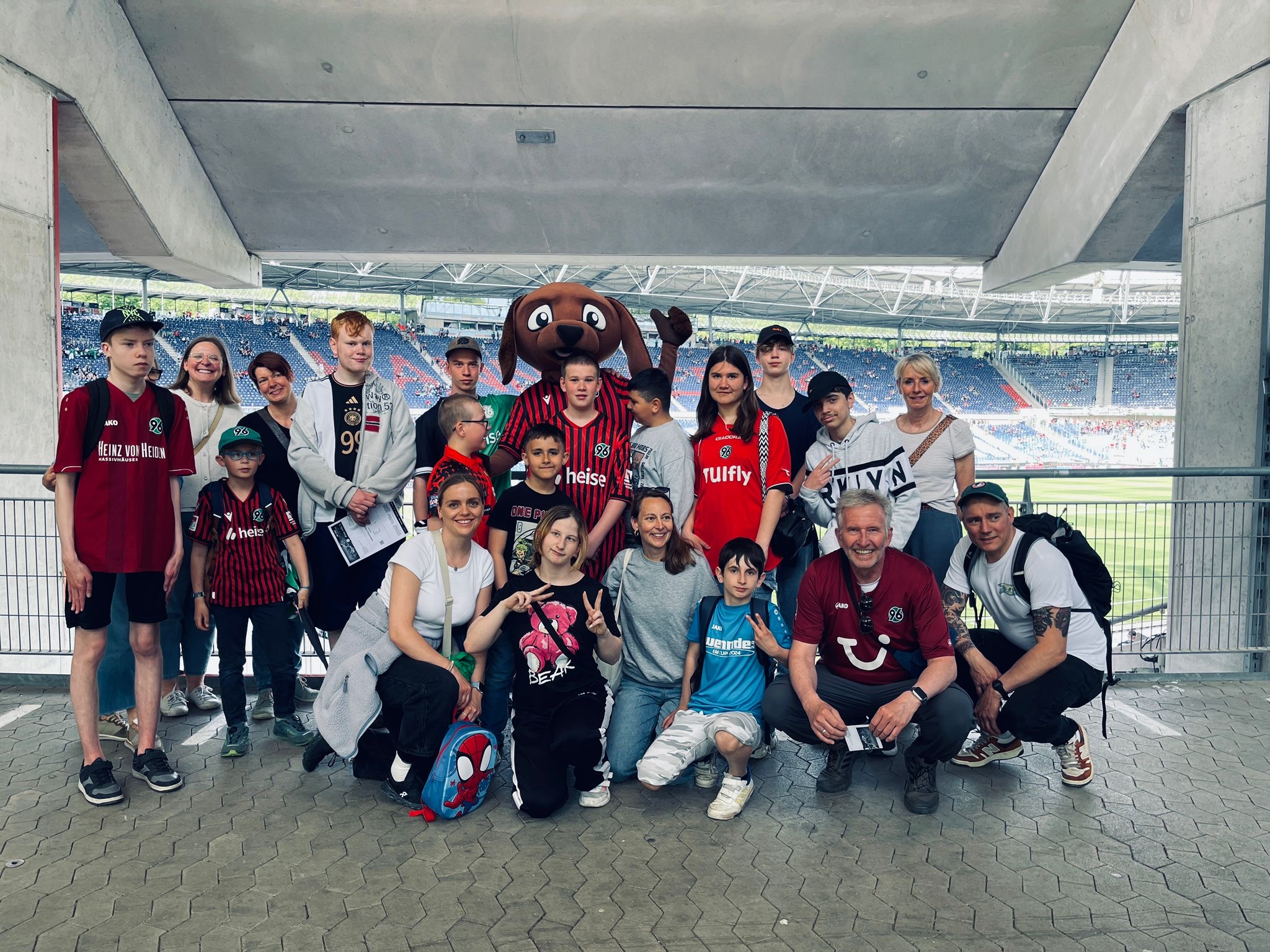 Eine Gruppe von Menschen, darunter Kinder und Erwachsene, posieren gemeinsam in einem Stadion. Einige tragen Fußballtrikots und eine Person in einem großen braunen Maskottchenkostüm steht im Hintergrund. Das Spielfeld und die Tribünen sind im Hintergrund zu sehen.