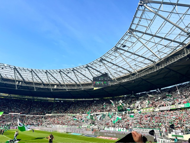 Ein großes Stadion mit offenem Dach, gefüllt mit jubelnden Fans, die grüne und weiße Fahnen schwenken, unter einem klaren blauen Himmel. Die Tribünen sind voll, und die moderne Struktur des Stadions ist über dem Kopf zu sehen.